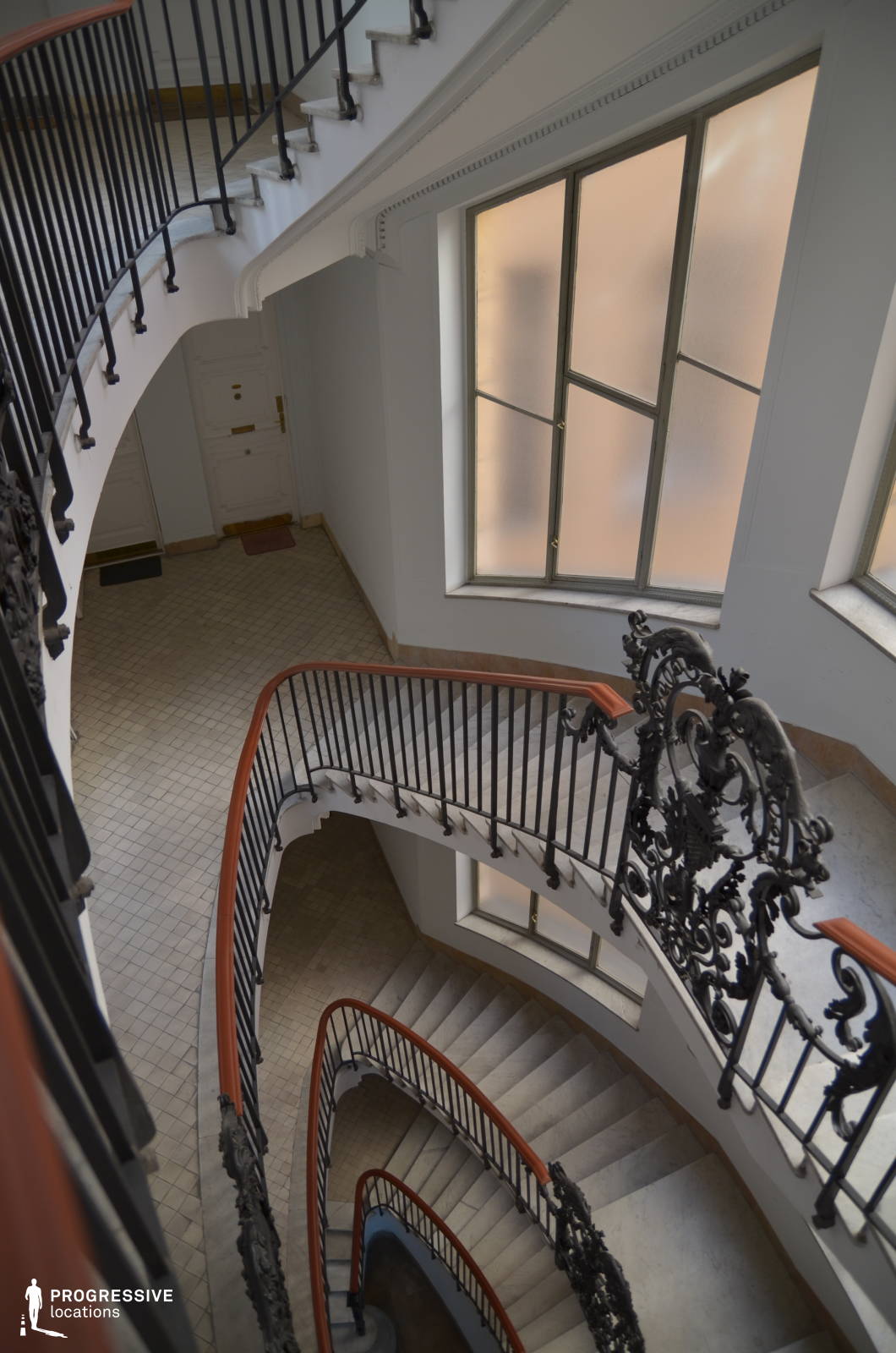 This sophisticated oval staircase winds gracefully through a light-filled atrium, framed by sleek black wrought-iron railings with intricate decorative flourishes. The white marble steps contrast with the warm wooden handrails, while frosted glass windows diffuse soft natural light, enhancing the airy and refined atmosphere. The flowing curves of the stairwell create a dynamic sense of movement and depth, making this location ideal for period dramas, psychological thrillers, or high-society intrigue films. Its combination of classical elegance and minimalist sophistication provides a versatile and visually compelling backdrop for cinematic storytelling.