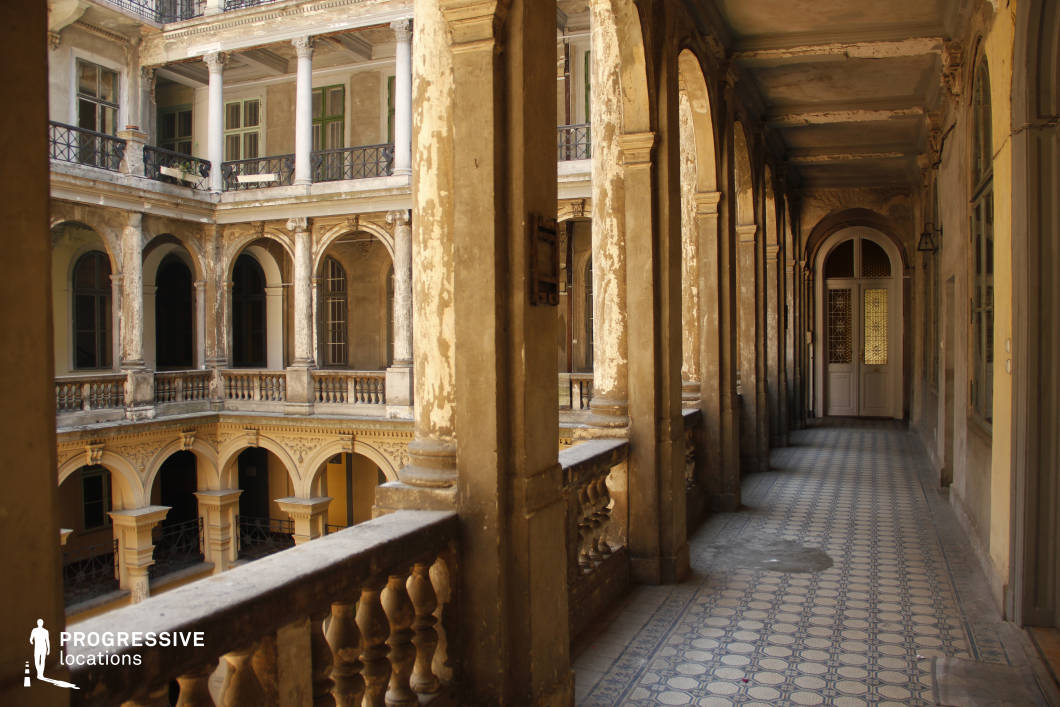 This image captures a historic, multi-level courtyard with a series of elegant arches and classical columns that frame the space with a sense of faded grandeur. The peeling plaster, weathered surfaces, and intricate balustrades emphasize the building’s aged character, while the patterned tiled walkway adds a refined yet deteriorated aesthetic. The layered architecture, featuring arched openings, decorative moldings, and wrought iron railings, creates depth and visual intrigue. This location is ideal for filming period dramas, gothic thrillers, or historical mystery films, offering a rich, atmospheric setting that evokes nostalgia, elegance, and a sense of lost opulence.