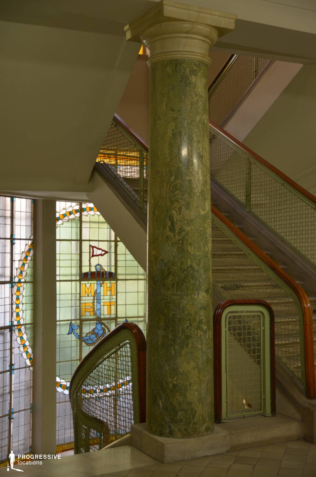 A vintage staircase featuring an elegant green marble column, metal railings with intricate mesh detailing, and polished wooden handrails. The background is dominated by a stunning stained-glass window, displaying nautical-themed designs and the initials 