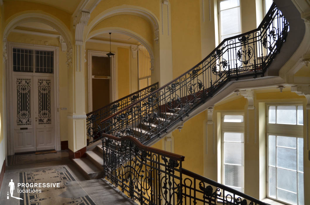 The image showcases a grand, historic interior featuring an ornate staircase with intricate wrought iron railings adorned with floral motifs. The staircase has a sweeping, curved design with a polished wooden handrail, enhancing its refined aesthetic. The surrounding architecture includes tall, arched doorways, decorative moldings, and soft yellow walls, which contrast with the dark metalwork and add warmth to the scene. Large windows allow natural light to filter in, casting subtle shadows that highlight the space's depth and textures. The patterned tile flooring and intricate door details contribute to the vintage elegance of the location. This setting is ideal for filming period dramas, romantic narratives, or sophisticated mystery films, offering a visually rich backdrop that evokes history, refinement, and intrigue.