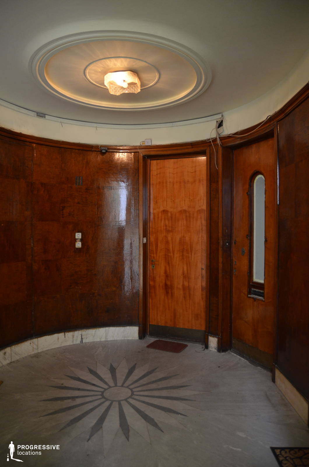 This unique circular entrance hall features dark, polished wood paneling that curves seamlessly around the space, giving it a refined yet slightly mysterious ambiance. The floor is adorned with a striking radial marble inlay design, adding an element of symmetry and elegance. A simple yet stylish ceiling light fixture sits within a concentric circular molding, enhancing the mid-century aesthetic. The doors, each with distinct detailing—one featuring a narrow, arched window—add to the intrigue of the space. This setting is well-suited for film genres such as period dramas, psychological thrillers, or noir-inspired mysteries, offering a sophisticated and slightly enigmatic atmosphere.