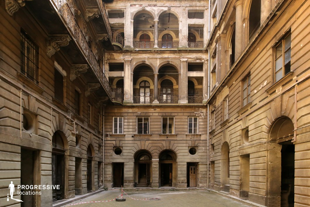 This weathered neoclassical courtyard features grand yet deteriorating facades, adorned with layered archways, fluted columns, and ornate iron railings. The aged stone surfaces and peeling plaster reveal the building’s historical significance, while the worn-out architectural details add to its hauntingly atmospheric appeal. The multi-story balconies and open corridors create a strong sense of depth, making the space feel both imposing and intimate. This location is ideal for gothic dramas, historical thrillers, or post-apocalyptic films, offering a visually rich and immersive setting that conveys themes of mystery, decay, and forgotten grandeur.