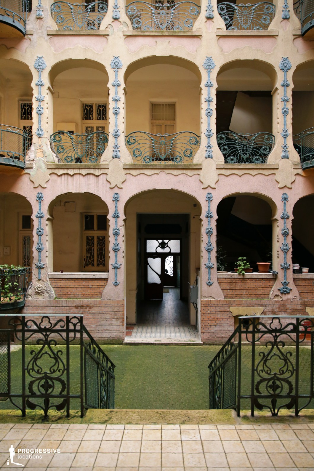 This enclosed courtyard showcases an exquisite Art Nouveau design, featuring soft pastel-toned walls adorned with delicate blue floral embellishments and elegantly curved wrought-iron balconies. The arched openings and intricate detailing on the railings emphasize a refined, organic aesthetic. The central staircase and entranceway, framed by decorative metalwork, add depth to the composition, leading toward an inviting passage lined with patterned tiles. The aged surfaces and gentle color palette contribute to a dreamlike, vintage atmosphere. This visually rich setting is perfect for romantic period films, historical dramas, or whimsical fantasy productions, offering a unique and sophisticated backdrop.