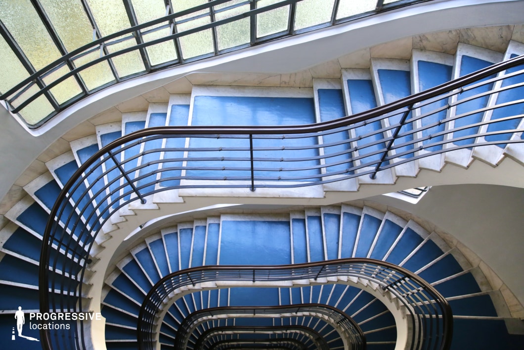 This dynamic, multi-level staircase showcases a sleek modernist design with bold blue steps framed by smooth white marble edges. The flowing curvature of the stairwell is accentuated by streamlined metal railings that follow its organic form. Large frosted glass windows line the upper level, allowing diffused natural light to cast soft reflections on the surfaces, enhancing the architectural depth. The combination of cool-toned colors and geometric repetition creates a visually striking effect, making this location ideal for artistic cinematography, contemporary thrillers, or sci-fi narratives. Its clean design and dramatic spiraling perspective offer excellent opportunities for visually engaging storytelling.