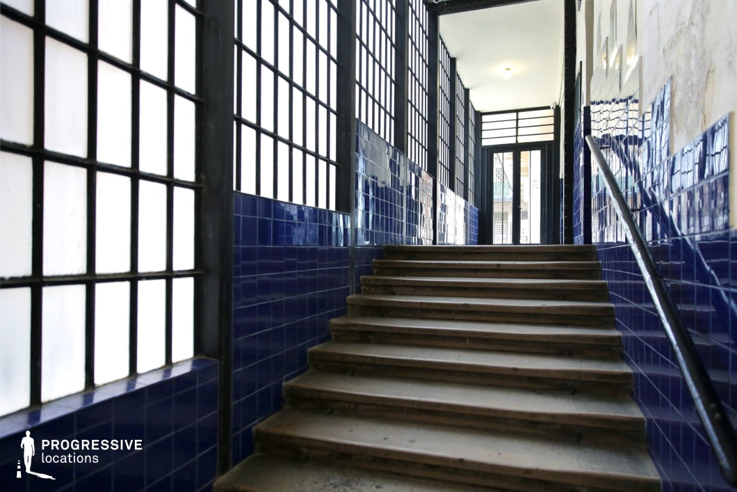 This narrow, industrial-style stairway is defined by its deep blue ceramic-tiled walls and large grid-patterned frosted windows, which allow soft, diffused light to filter into the space. The worn stone steps and black metal handrail emphasize a utilitarian, early 20th-century aesthetic, while the interplay of textures—smooth tiles, rough concrete, and structured windowpanes—adds visual contrast. The perspective leads towards a glass-paneled door at the top, reinforcing a sense of depth and transition. The stark yet atmospheric setting makes this location ideal for noir films, urban dramas, or psychological thrillers, offering a cinematic blend of mystery and raw architectural character.