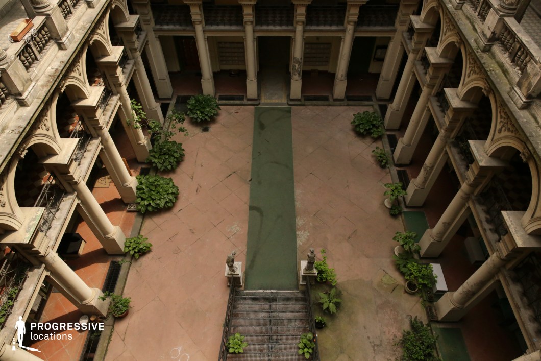 This expansive courtyard, viewed from above, is framed by a series of elegant neoclassical arches and columns, lending a sense of grandeur and historical depth. The worn stone flooring, intersected by a faded green pathway, creates a striking visual contrast with the lush greenery placed along the edges. A symmetrical staircase flanked by statues leads into the space, reinforcing the classical aesthetic. The aging facade, softened by creeping vegetation and weathered textures, adds an atmospheric charm. This location is ideal for period dramas, historical thrillers, or mystery films, offering a rich and immersive setting for storytelling.