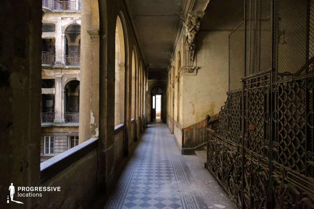 This atmospheric neoclassical corridor exudes a sense of faded grandeur, with a long, arched walkway flanked by weathered stone columns and an intricate wrought-iron elevator cage. The ornate details, including sculptural reliefs and patterned tile flooring, contrast with the peeling walls and aged textures, creating a visually rich setting filled with history. The perspective draws the eye toward the distant doorway, enhancing the sense of depth and mystery. This location is ideal for period dramas, gothic thrillers, or noir-inspired films, offering a hauntingly beautiful backdrop that evokes a forgotten era of elegance and intrigue.
