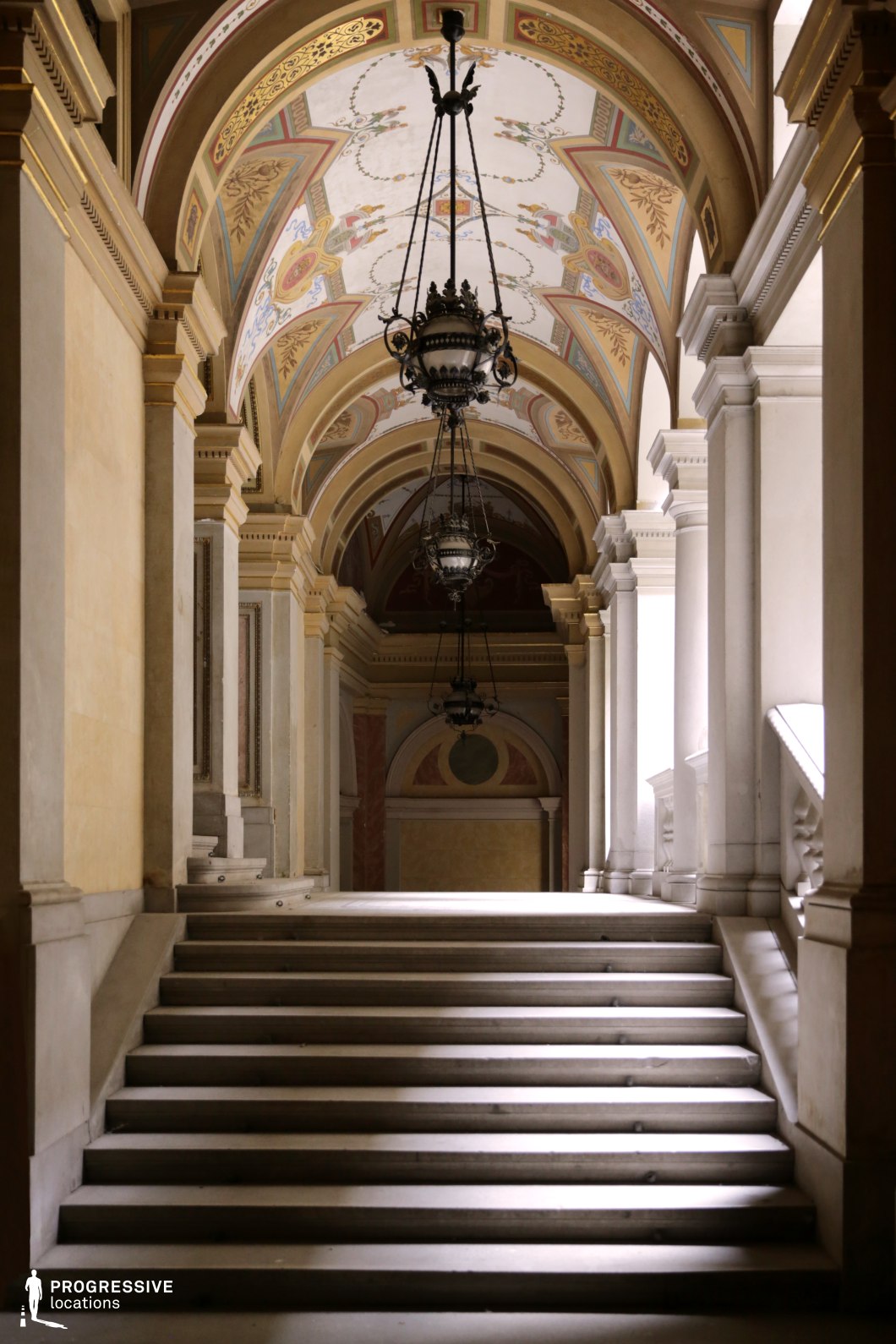 This magnificent neoclassical staircase ascends beneath a series of richly decorated arches adorned with intricate frescoes, gold detailing, and sculptural moldings. Suspended lantern-style chandeliers enhance the regal atmosphere, while towering white columns frame the space with a sense of grandeur. The play of light and shadow across the stone steps and balustrades adds depth, emphasizing the architectural precision. This setting is ideal for period dramas, historical epics, or aristocratic intrigue films, offering a visually stunning and immersive location that exudes elegance, power, and sophistication.