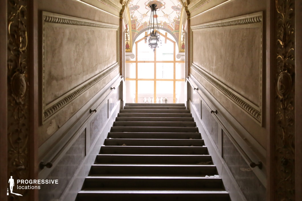 This majestic staircase ascends toward a luminous arched window, framed by intricate gold-trimmed moldings and classical paneling. The walls feature finely carved decorative elements, enhancing the space’s opulent character. A grand chandelier hangs from the elaborately painted ceiling, adding to the regal atmosphere. The worn stone steps and subtle signs of aging contrast with the elegance of the surrounding architecture, reinforcing a sense of history and faded grandeur. This location is ideal for filming period dramas, historical intrigue, or gothic thrillers, offering a visually rich and atmospheric setting that evokes sophistication and timeless allure.