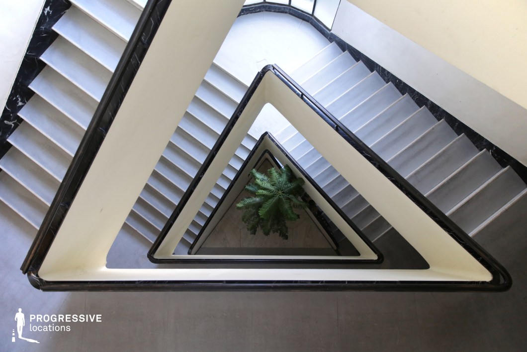 This striking staircase showcases a rare triangular design, with sharp angular lines and contrasting black marble railings against smooth white walls. The repeating geometric pattern creates a mesmerizing visual rhythm, drawing the eye downward to the central atrium where a lush green plant softens the rigid architectural elements. The clean, minimalist aesthetic is complemented by natural lighting and sleek stair treads, reinforcing the modernist style. This location is ideal for filming psychological thrillers, neo-noir dramas, or avant-garde productions, offering a dynamic and unconventional setting that plays with perspective and depth.