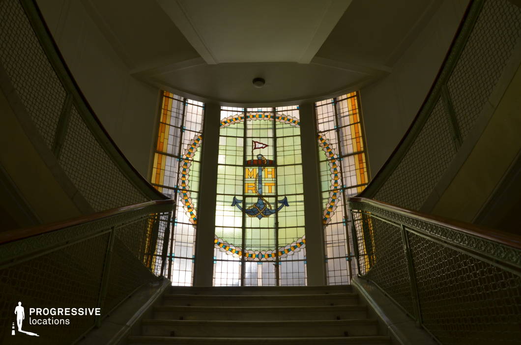 This elegant staircase leads up to a striking stained glass window, which serves as the architectural focal point. The window features an intricate circular design with nautical elements, framed by warm amber and soft green hues that diffuse natural light into the space. The stair railings are adorned with decorative metalwork, complementing the vintage aesthetic. The symmetrical composition and upward perspective add a sense of grandeur and purpose. This location is ideal for filming historical dramas, period mysteries, or atmospheric noir films, offering a rich visual backdrop that conveys heritage, prestige, and a timeless elegance.