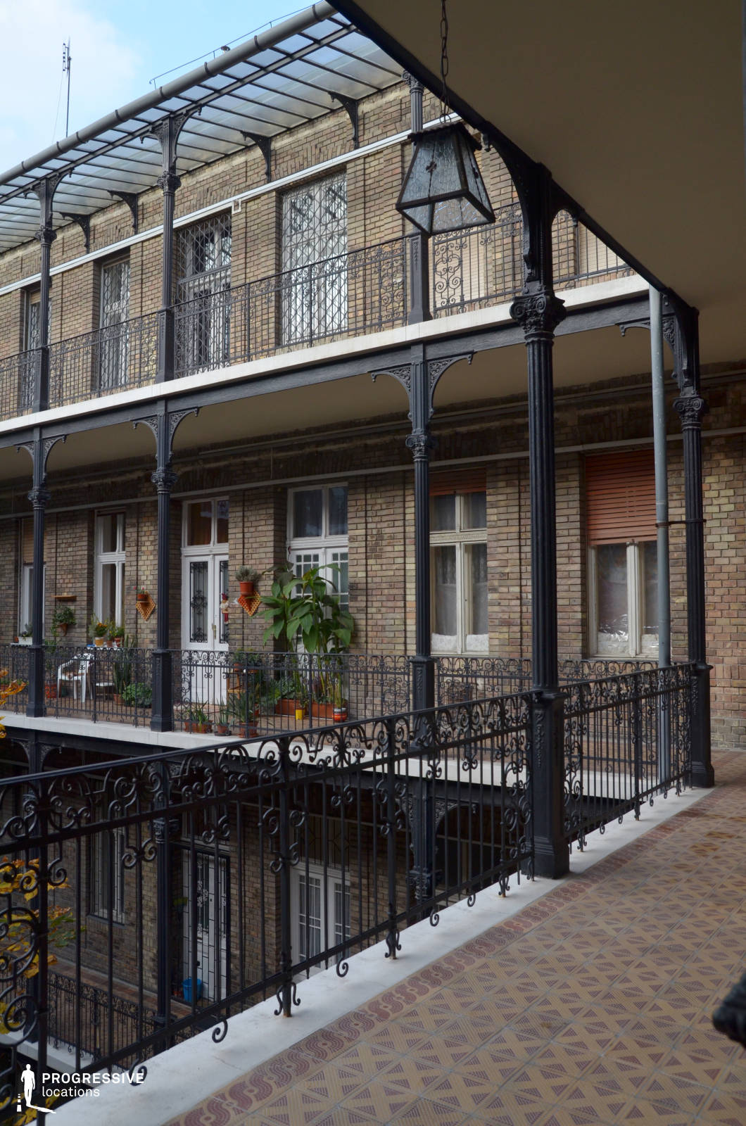 This elegant courtyard features intricate cast-iron railings and columns that support a covered walkway, giving the space a refined, vintage charm. The brick façade of the surrounding building adds warmth and texture, while decorative window grilles enhance the historical character. The patterned tile flooring and delicate metalwork on the balconies evoke a late 19th or early 20th-century aesthetic. The open yet intimate layout allows for dynamic visual compositions, making this location ideal for period dramas, romantic narratives, or films exploring urban nostalgia and hidden architectural gems.
