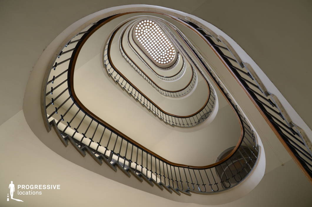 This visually striking spiral staircase features smooth, continuous curves that ascend in a hypnotic, elliptical pattern. The clean white walls contrast with the sleek black metal railings and polished wooden handrails, emphasizing the modernist design. The repetition of levels and the perforated ceiling skylight create a sense of infinite depth and symmetry, making this location ideal for dynamic camera movements. The elegant, minimalist aesthetic suits psychological thrillers, avant-garde films, or high-concept sci-fi productions, offering a mesmerizing backdrop that plays with perception and movement.