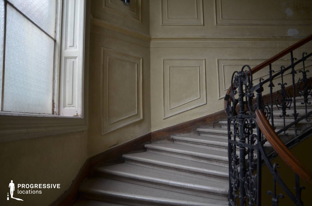 This elegantly aged staircase features a beautifully detailed wrought iron railing with intricate scrollwork, topped with a polished wooden handrail. The softly worn stone steps curve gently upward, framed by classic paneled walls with subtle decorative molding. A large frosted window allows diffused light to highlight the textures of the space, casting gentle shadows that enhance the depth and historic ambiance. The muted color palette of beige and cream tones adds to the timeless character. This location is ideal for filming period dramas, gothic thrillers, or mystery narratives, offering a refined yet slightly melancholic atmosphere.
