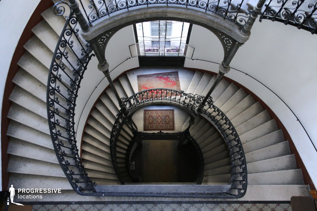 This striking, curved stone staircase spirals downward in a seamless, flowing motion, framed by intricate wrought-iron railings that add a sense of grandeur and craftsmanship. The perspective from above enhances the depth and symmetry of the design, drawing the eye toward the lower level, where patterned rugs and an open window introduce warmth and contrast. The soft natural light filtering through the window highlights the texture of the stone steps and the elegant curvature of the balustrade. This visually dramatic location is ideal for period films, mystery thrillers, and psychological dramas, offering a cinematic backdrop for suspenseful or introspective scenes.