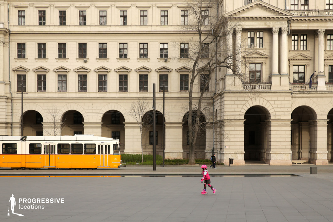 A grand neoclassical-style building with symmetrical facades, arched walkways, and intricate detailing dominates this urban scene. The uniform cream-colored exterior, decorative columns, and evenly spaced windows create a stately and elegant backdrop. A yellow tram, characteristic of European public transportation, adds a dynamic element, reinforcing the city's historical yet functional infrastructure. The expansive, open square in the foreground, paved with large, smooth tiles, offers a modern contrast to the traditional architecture. The linear and geometric composition, along with the interplay of classic and contemporary elements, makes this location ideal for cinematic or commercial projects requiring a sophisticated yet authentic European cityscape.