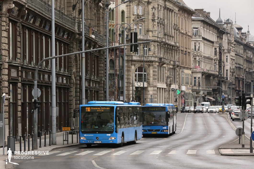 A visually rich urban setting where historic, ornately decorated buildings line a curved city street. The facades feature intricate stonework, wrought-iron balconies, and a mix of architectural styles, creating a timeless and elegant backdrop. The presence of blue public buses in motion highlights the city's efficient transportation system, adding a contemporary, functional contrast to the old-world architecture. The wide road, flanked by modern street elements like traffic lights, pedestrian crossings, and lampposts, enhances the metropolitan atmosphere. This location is ideal for capturing a European city’s blend of historic grandeur and modern-day functionality, making it suitable for film scenes emphasizing urban life, cultural richness, or architectural storytelling.