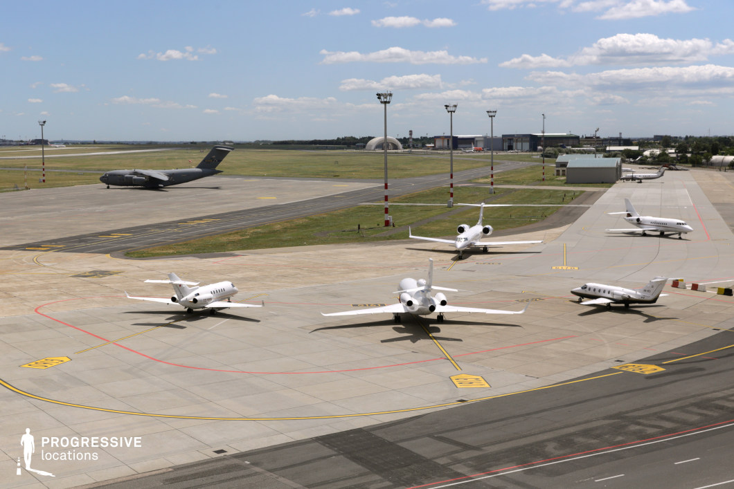 A well-organized airport apron scene featuring multiple private jets parked in a structured formation, with a military cargo aircraft taxiing in the background. The airfield extends into the distance, lined with hangars, service buildings, and red-and-white striped light poles, emphasizing the operational scale of the location. The contrast between the sleek, white private jets and the imposing, dark-colored military plane adds a layered visual dynamic. The vast concrete surfaces, marked with taxi lines and directional signage, contribute to a highly structured and functional aesthetic. This setting is ideal for aviation-related cinematic or commercial projects, offering a clean, professional atmosphere with both civilian and military aviation elements.