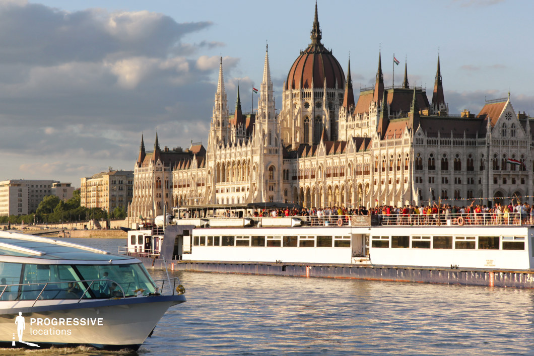 A grand architectural scene featuring the neo-Gothic Hungarian Parliament Building along the riverbank, its intricate spires and detailed facade standing as a striking focal point. In the foreground, river cruise boats glide across the water, enhancing the scene’s lively, touristic atmosphere. The contrast between the modern glass-roofed boat and the larger, classic-style cruise ship filled with passengers creates a dynamic visual composition. The rippling water reflects the historic architecture, adding depth and movement to the image. This location is ideal for cinematic and commercial projects showcasing European elegance, cultural richness, or luxury river tourism.