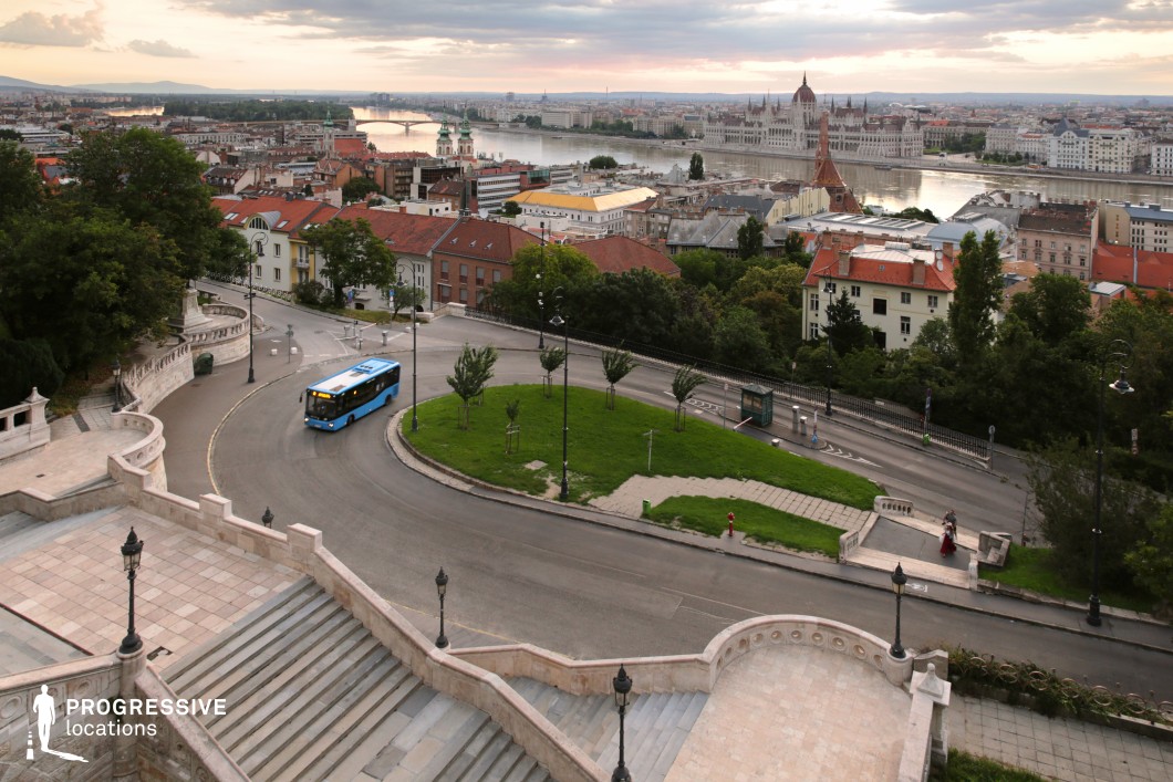 A breathtaking elevated view of a historic European city, with the iconic Parliament Building and the Danube River serving as the focal points in the background. The foreground features an elegant stone staircase with intricate balustrades, leading down to a curved roadway lined with vintage-style street lamps and lush greenery. A blue public bus navigates the winding street, adding a touch of modern urban life to the otherwise classical setting. The red rooftops, historic churches, and mix of architectural styles create a rich, visually dynamic cityscape. This location offers a perfect blend of historic grandeur and everyday city movement, making it ideal for cinematic and commercial projects seeking a picturesque yet authentic European atmosphere.