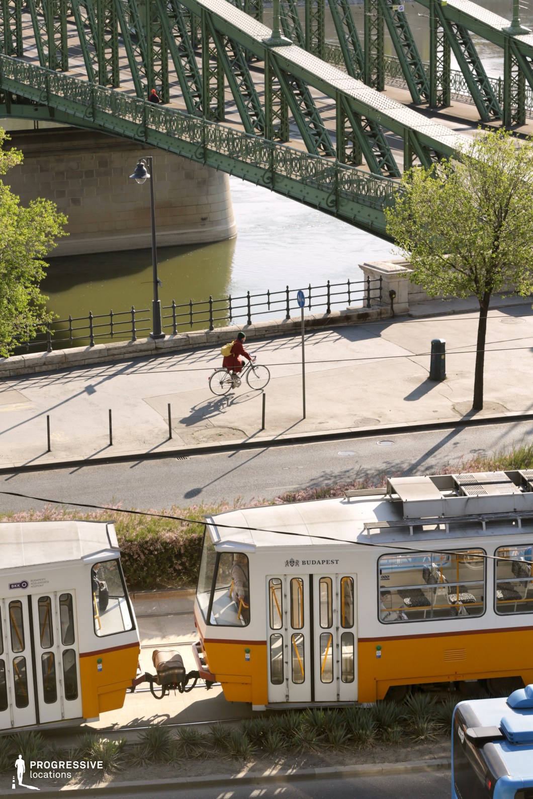 A vibrant urban scene featuring a classic green steel bridge with intricate metalwork spanning over a calm river, creating a strong architectural focal point. Below the bridge, a pedestrian and cycling path runs parallel to the water, enhancing the location’s accessibility and movement. In the foreground, two yellow trams with their vintage-inspired design add an element of dynamic urban transport. The scene is framed by lush greenery and cast shadows, contributing to a balanced composition that blends historic infrastructure with everyday city life. This setting is ideal for cinematic and commercial projects seeking a timeless European cityscape with a rich architectural and transport heritage.