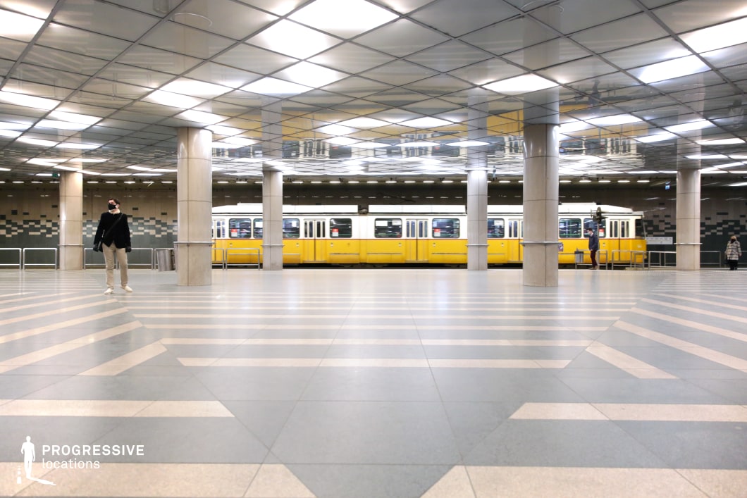 A striking underground transport hub with a highly structured geometric design, featuring reflective ceiling panels, patterned flooring, and robust concrete pillars. The symmetry and repetition of the architectural elements create a futuristic and modern visual appeal. A classic yellow tram is stationed in the background, adding a nostalgic contrast to the sleek, contemporary setting. The expansive open space, marked by bold diagonal floor patterns, enhances the depth and perspective of the composition. This location is ideal for cinematic projects requiring a clean, structured urban backdrop with a mix of historical and modern transportation aesthetics.
