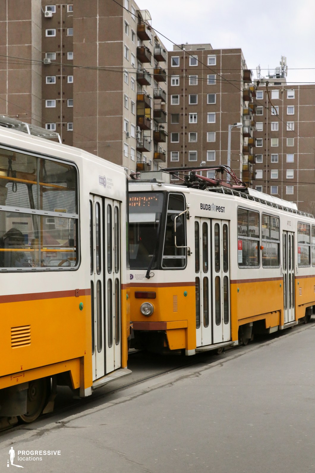 A dense urban scene featuring two iconic yellow trams navigating a residential area dominated by large, uniform socialist-era apartment buildings. The tram’s vintage design, with its tall windows and curved edges, contrasts with the repetitive geometric patterns of the surrounding architecture. Overhead electric cables crisscross above, reinforcing the structured and functional aesthetic of the setting. The muted tones of the apartment blocks enhance the visual prominence of the trams, making them the focal point of the image. This location is ideal for cinematic projects seeking an authentic Eastern European atmosphere, emphasizing urban life, historical public transport, or a post-socialist cityscape.