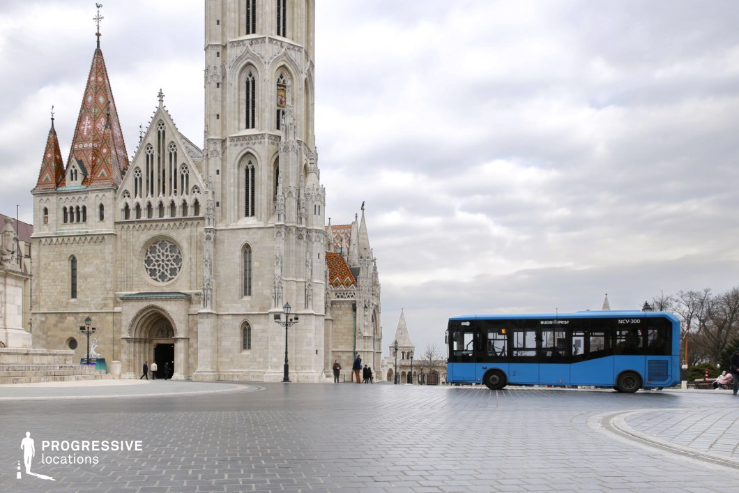 A striking juxtaposition of historical and contemporary elements, featuring a grand Gothic-style cathedral with intricate stonework, towering spires, and colorful tiled roofs. The detailed architecture of the church, with pointed arches and elaborate carvings, serves as a dominant focal point. In contrast, a sleek blue city bus navigates the cobblestone plaza, integrating modern urban mobility into this timeless setting. The curved road, framed by historic lampposts and stone balustrades, adds depth and movement to the composition. This location is ideal for cinematic and commercial projects that highlight the fusion of historic European architecture with modern city infrastructure.