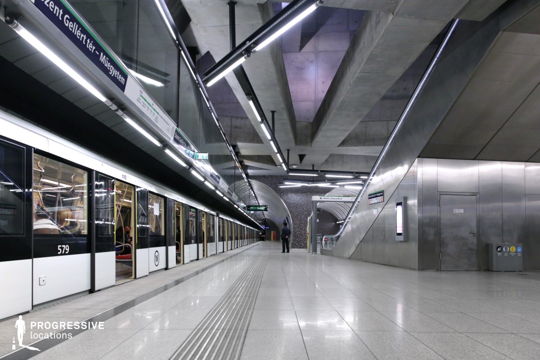 A strikingly modern metro station characterized by bold geometric concrete structures, polished metallic surfaces, and linear lighting that enhances the futuristic aesthetic. The platform is spacious and minimalistic, with a sleek metro train stopped alongside, featuring large glass windows and a monochromatic color scheme. Overhead, angular concrete beams create a dramatic sense of depth, complemented by reflective tiled flooring that amplifies the station's symmetry. The escalators and signage contribute to the high-tech, efficient urban infrastructure. This location is ideal for cinematic or commercial projects requiring a sleek, industrial, or sci-fi-inspired underground transit setting.