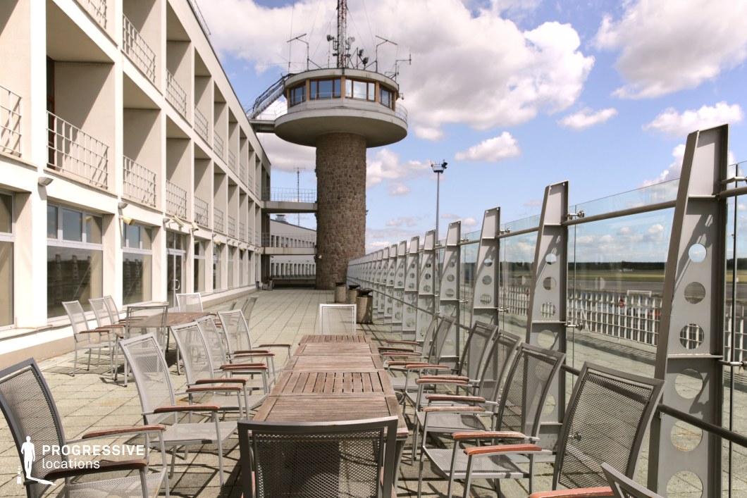 A unique aviation-themed location featuring an expansive outdoor terrace overlooking an airport runway. The setting includes a long wooden table surrounded by metal-framed chairs, suggesting a space designed for casual gatherings or observation. A prominent control tower with a circular glass observation deck, supported by a cylindrical stone base, stands as a focal point, adding an industrial yet functional element to the scene. The adjacent building’s repetitive grid-like facade, with uniform balconies and large windows, enhances the structured aesthetic. The terrace is enclosed by a modern glass railing with metallic supports, providing an unobstructed view of the airfield. This location is ideal for cinematic and commercial projects requiring an aviation-related backdrop with a blend of retro and contemporary architectural influences.
