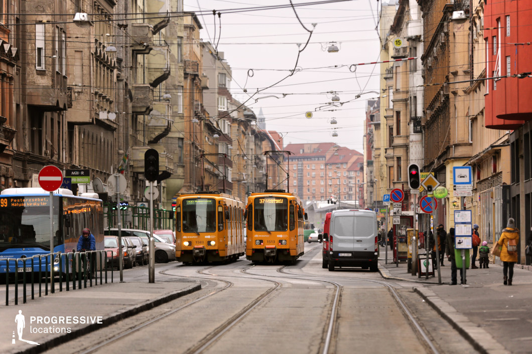 A dense, dynamic city street framed by historic buildings with ornate facades, peeling plaster, and a mix of architectural styles, showcasing a rich urban texture. Overhead, a web of tram cables crisscrosses the scene, emphasizing the well-integrated public transportation system. Two yellow trams navigate the curving tracks, their bright color standing out against the muted tones of the surrounding environment. The street is filled with varied elements, including a blue city bus, parked cars, bicycles, pedestrians, and a network of street signs and traffic lights, creating an authentic metropolitan atmosphere. The perspective leads the eye toward the distant, more modern red-roofed buildings, adding depth and contrast. This location is ideal for cinematic and commercial projects that require a raw, energetic European cityscape blending historical charm with contemporary urban life.
