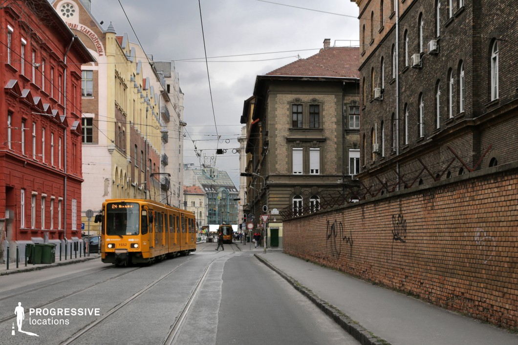 A visually rich city street lined with diverse architectural styles, featuring a mix of vibrant and muted facades. The bold red and yellow buildings contrast sharply with the dark brick structures, adding depth and texture to the scene. A classic yellow tram moves along curved tracks, its bright color standing out against the subdued tones of the surrounding environment. The aged brick wall in the foreground, topped with metal barriers, introduces an element of urban grit, while graffiti adds to the street’s character. Overhead tram cables crisscross the sky, reinforcing the structured transportation network. This location is ideal for cinematic and commercial projects requiring an old European cityscape with a mix of historic charm and raw urban energy.