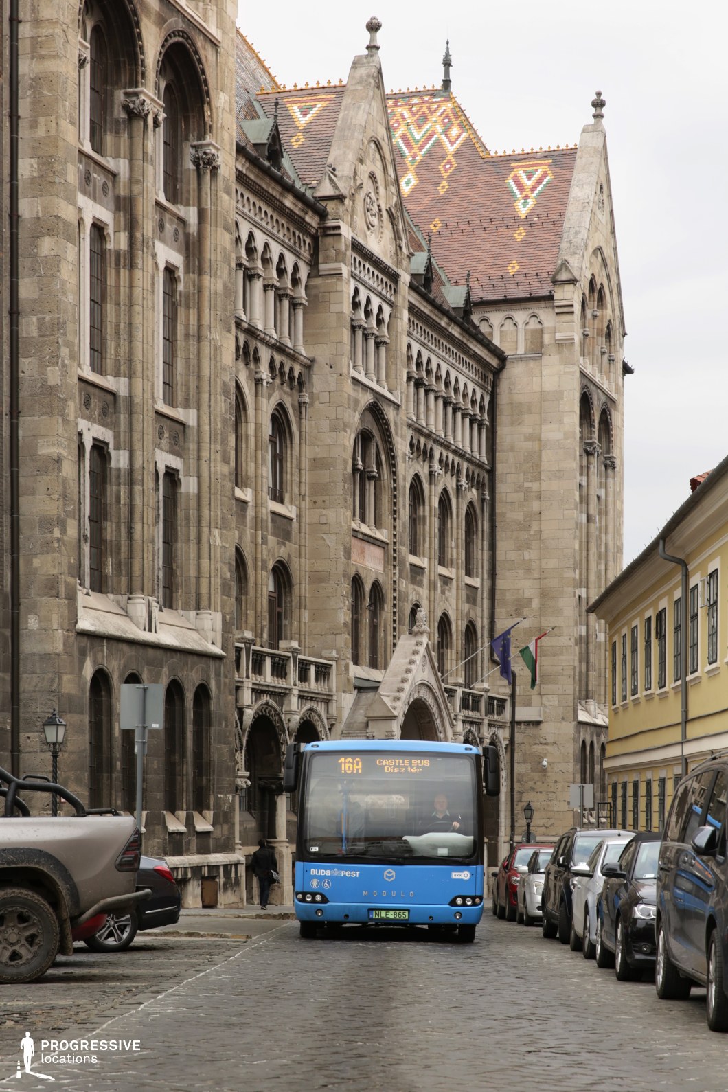 A grand, historic street framed by monumental Gothic-style architecture, featuring detailed stone facades, arched windows, and intricate ornamentation. The colorful, patterned tiled roof adds a striking visual contrast to the otherwise neutral-toned stonework. A small blue city bus navigates the narrow cobblestone street, highlighting the integration of modern public transportation within a centuries-old urban setting. The composition is enriched by the juxtaposition of parked cars, historic lampposts, and flags, creating a dynamic blend of heritage and contemporary functionality. This location is ideal for cinematic and commercial projects seeking an authentic European old-town atmosphere with a unique contrast between medieval architecture and modern infrastructure.