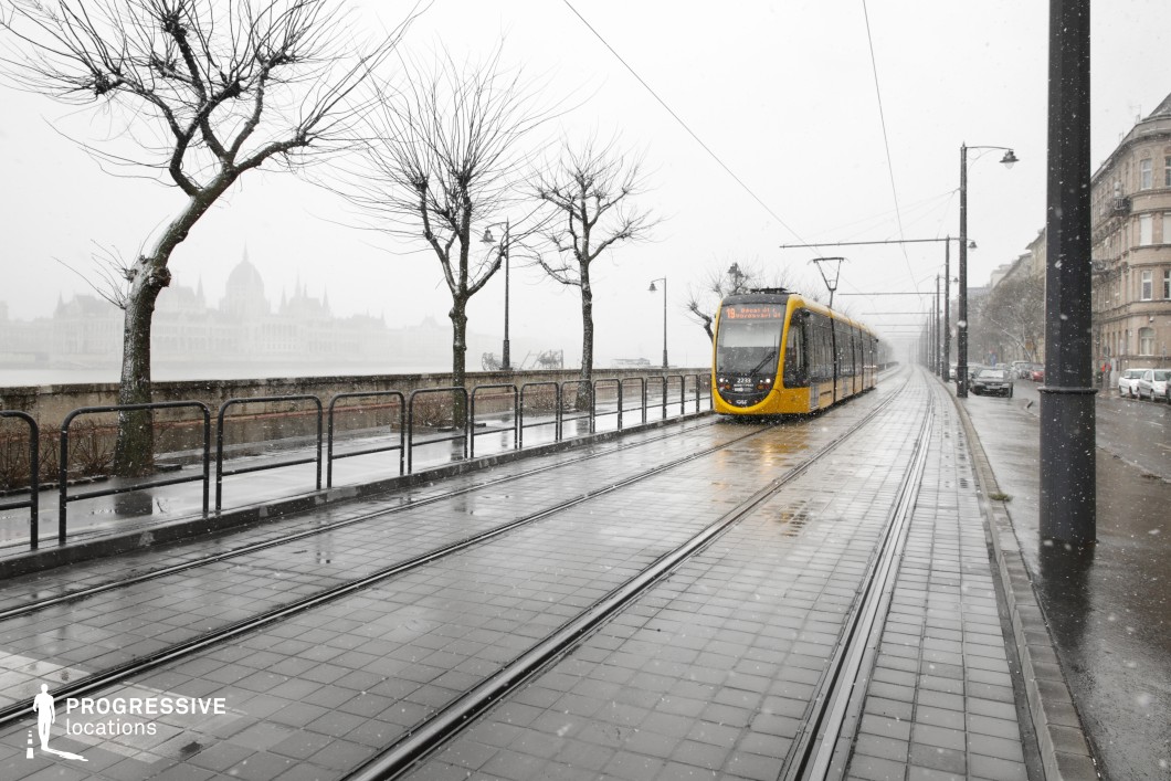 A sleek modern yellow tram travels along a scenic riverside tramline, bordered by elegant historic buildings and lined with leafless trees. The tracks follow the curve of the riverbank, adding depth and perspective to the composition. Overhead tram cables crisscross the sky, reinforcing the structured urban infrastructure. The subdued tones of the environment contrast with the tram's bright yellow, making it a focal point in the scene. The distant silhouette of an iconic architectural landmark adds a timeless, atmospheric quality. This location is ideal for cinematic and commercial projects requiring a sophisticated European cityscape with a mix of historic elegance and modern public transport integration.