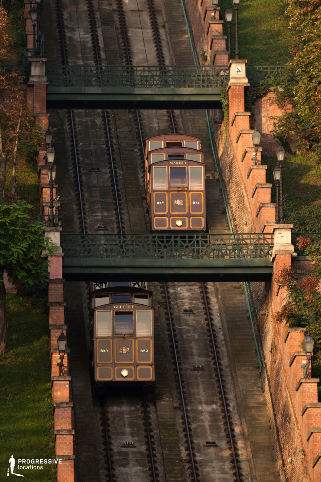 A beautifully preserved historic funicular railway ascends a steep hillside, flanked by elegant brick walls and vintage-style lampposts. The dark wooden cabins, adorned with gold detailing and traditional signage, exude old-world charm and sophistication. Two parallel tracks accommodate the counterbalanced funicular cars, passing under a decorative iron bridge that enhances the classic European aesthetic. The lush greenery surrounding the tracks, combined with the warm tones of the setting sun, adds depth and texture to the scene. This location is ideal for cinematic and commercial projects that require a timeless, historic city setting with a strong architectural and transportation heritage.