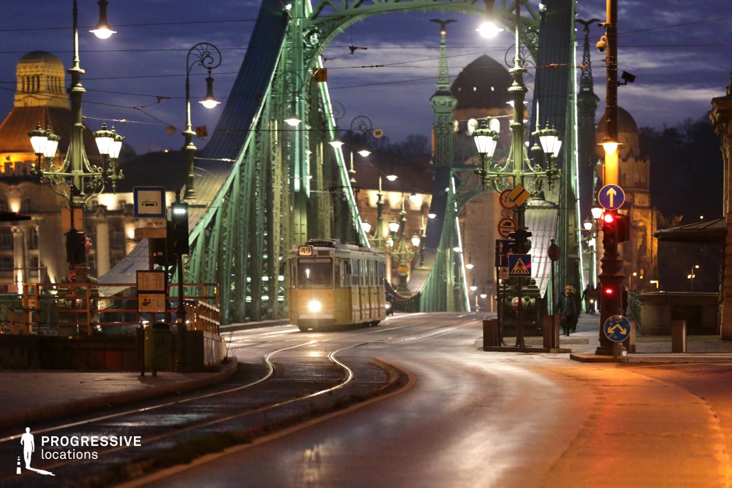 A stunning nighttime cityscape featuring a historic green iron bridge adorned with ornate lampposts, casting warm light onto the curving tram tracks. A classic yellow tram, its headlights glowing, moves across the bridge, adding a sense of urban motion and nostalgia. The intricate architectural details of the bridge, combined with the deep blue night sky and softly lit historic buildings in the background, create a cinematic and atmospheric setting. The winding street, reflective surfaces, and ambient streetlights enhance the depth and visual contrast. This location is ideal for cinematic and commercial projects requiring a moody, elegant European cityscape with a balance of history and modern urban life.