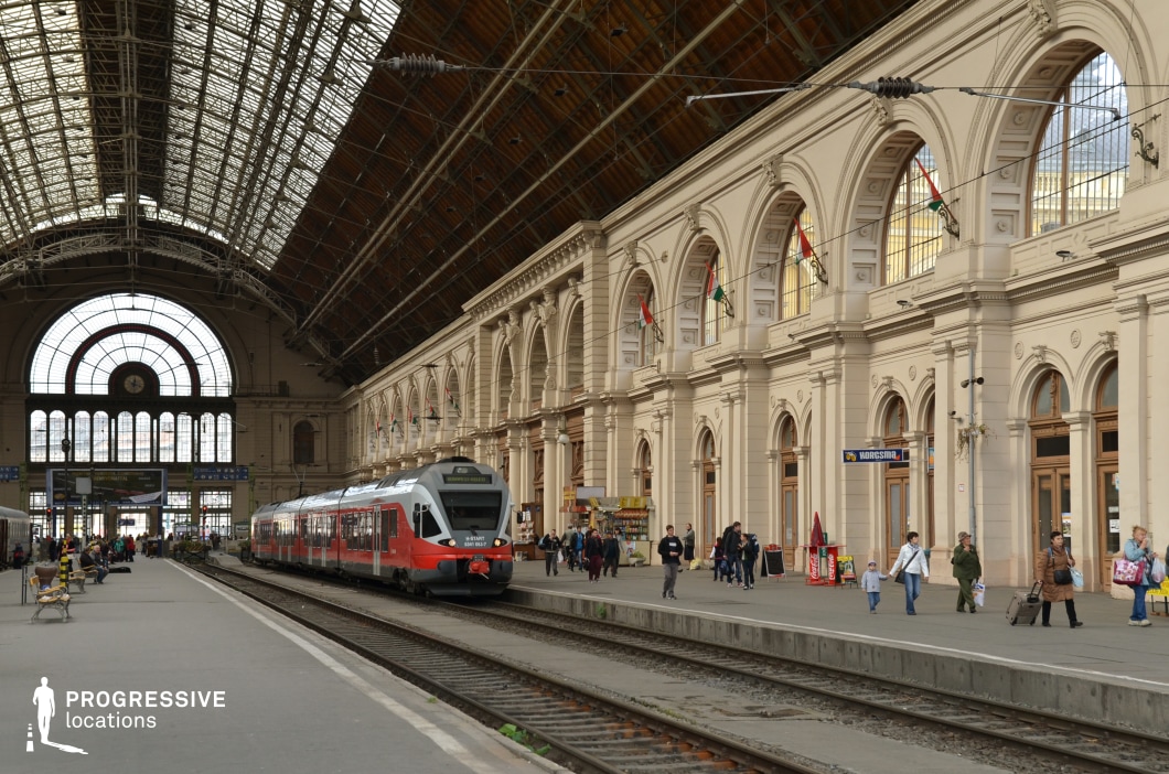 A magnificent European railway station with soaring iron-and-glass ceilings and an ornate neoclassical facade adorned with arched windows and decorative elements. The grand hall is bathed in natural light, highlighting its intricate details and creating an airy atmosphere. A sleek red and white modern train is stationed on the tracks, providing a striking contrast to the historic setting. Passengers move along the platform, adding life and dynamism to the scene. The large clock above the main entrance and the hanging signs contribute to the structured elegance of the station. This location is ideal for cinematic and commercial projects requiring a classic European train station setting, combining historic grandeur with contemporary transportation.