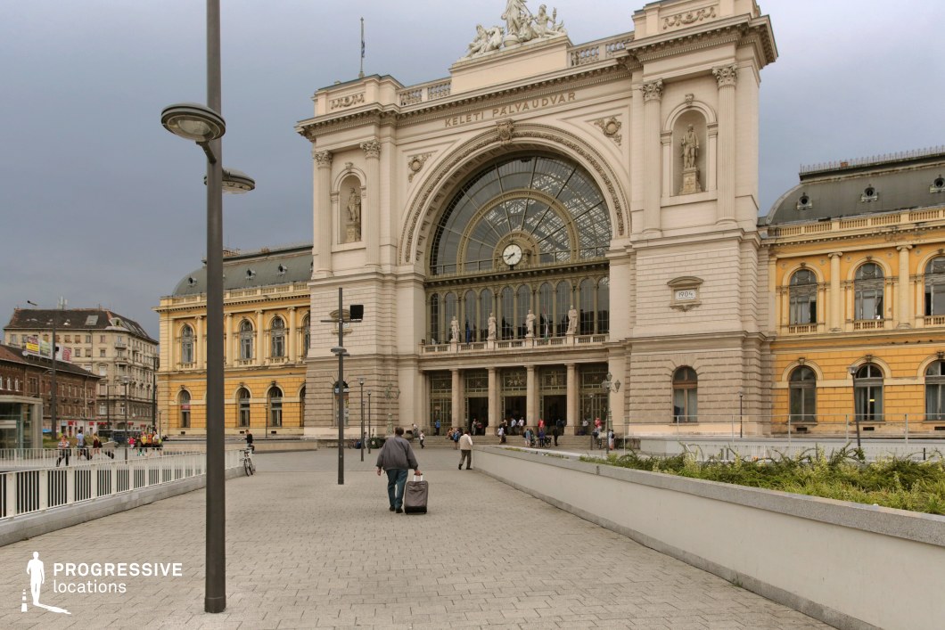A monumental railway station with a striking neoclassical facade, featuring an arched glass entrance, intricate sculptures, and a large central clock above the main doors. The building’s symmetrical design, with statues adorning the upper level, exudes grandeur and historic significance. The surrounding square is paved with modern stonework, leading toward the entrance where travelers with luggage and pedestrians move through the scene. The contrast between the richly decorated main station and the adjacent yellow-toned wing enhances the architectural depth. This location is ideal for cinematic and commercial projects requiring a grand European railway station setting, blending historic elegance with contemporary urban movement.