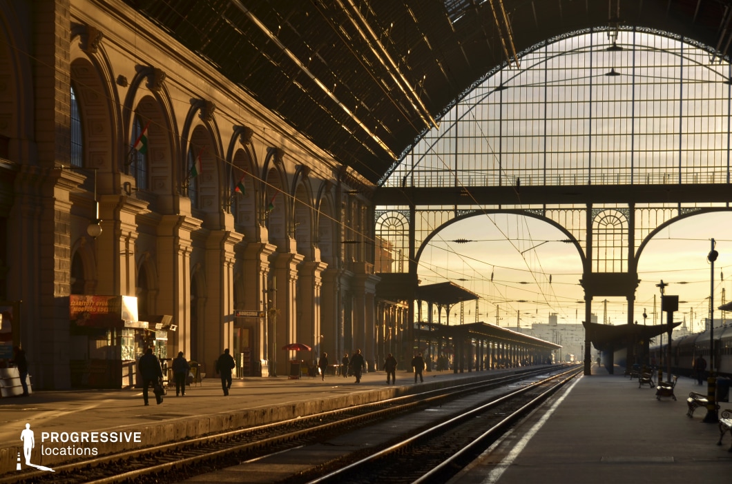 A historic European train station bathed in the golden light of sunrise, casting long shadows along the platform. The grand neoclassical facade with arched windows and intricate detailing is illuminated by the morning glow, emphasizing its architectural elegance. The massive glass-and-iron ceiling allows natural light to filter through, creating a warm and cinematic atmosphere. Passengers move through the space, adding a sense of life and travel anticipation. The railway tracks stretch into the distance, framed by elegant ironwork and towering columns. This location is ideal for film and commercial projects seeking a dramatic, historic train station with atmospheric lighting and a timeless European aesthetic.