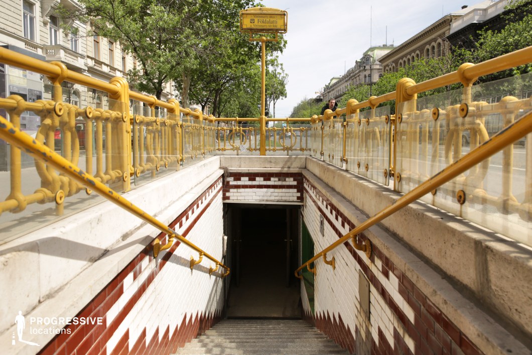 A charming and historic metro entrance with ornate yellow iron railings and glass panels, leading underground with tiled white and brown walls. The entrance has a vintage aesthetic, reminiscent of early 20th-century European design, blending classic craftsmanship with urban functionality. The descending staircase, framed by handrails, guides the viewer’s eye into the dimly lit passage below, creating a sense of depth and transition. In the background, elegant historic buildings and tree-lined streets add to the picturesque setting. This location is ideal for period-piece films, urban exploration themes, or projects requiring a visually rich and detailed metro access point with old-world charm.