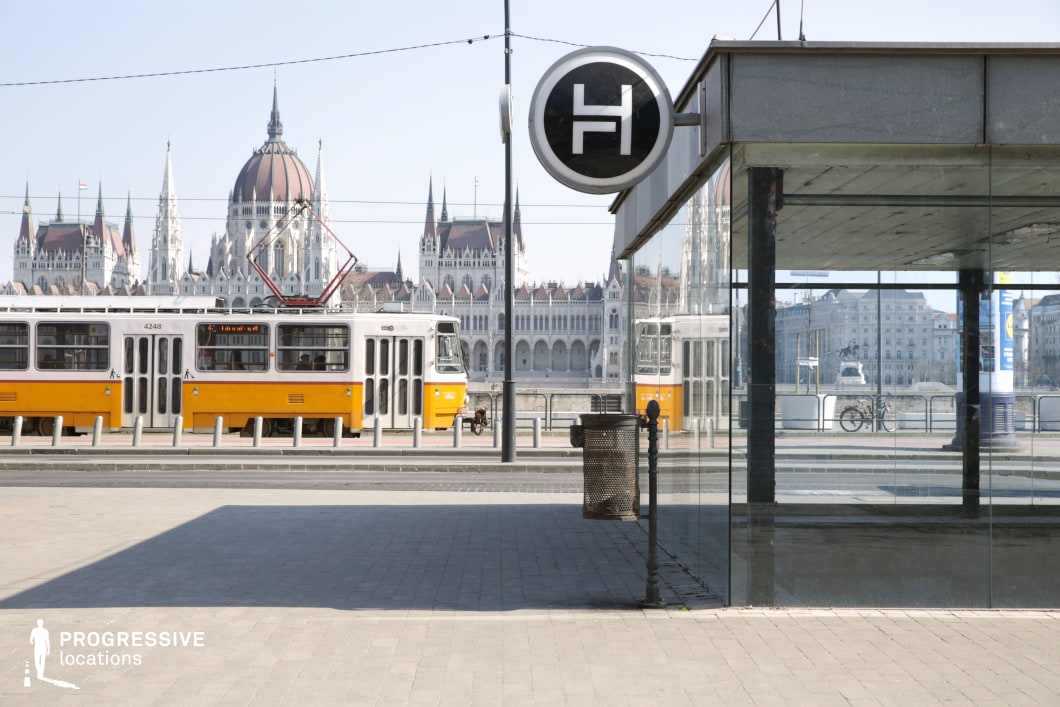 A striking urban scene featuring a modern tram stop with glass reflections, framing a classic yellow tram passing by. The imposing architecture of the Parliament building dominates the background, adding grandeur and a strong historical contrast to the contemporary glass structure in the foreground. The tram stop sign, with its minimalist circular design, blends seamlessly into the urban aesthetic. The clean lines, reflective surfaces, and symmetry of the image create a cinematic atmosphere, making this an excellent setting for transportation-related themes, city life narratives, or architectural contrasts in film and photography projects.
