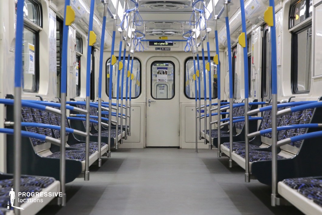 A clean and symmetrical metro train interior featuring blue and silver-toned grab bars and seats with a geometric pattern. The linear arrangement of poles and hanging straps creates a structured visual rhythm, while the soft lighting enhances the sense of space. Large windows allow for natural light during the day, while overhead digital displays provide real-time station updates. The interior's minimalistic and practical design makes it an ideal setting for scenes requiring a contemporary urban transit backdrop, such as commuter life, travel sequences, or cinematic explorations of public transportation.