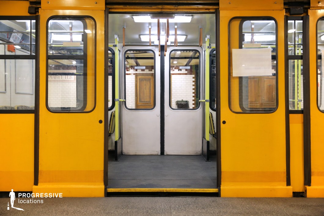 A vintage-style yellow metro train with its doors open at an underground station. The symmetrical composition highlights the doorway leading into the carriage, revealing a minimalist interior with wooden hand straps and muted colors. The exterior features large windows with black trim, providing reflections of the surrounding tiled walls with decorative brown accents. The retro aesthetic, combined with the warm artificial lighting, makes this an ideal location for scenes set in historical, nostalgic, or everyday urban transit settings.