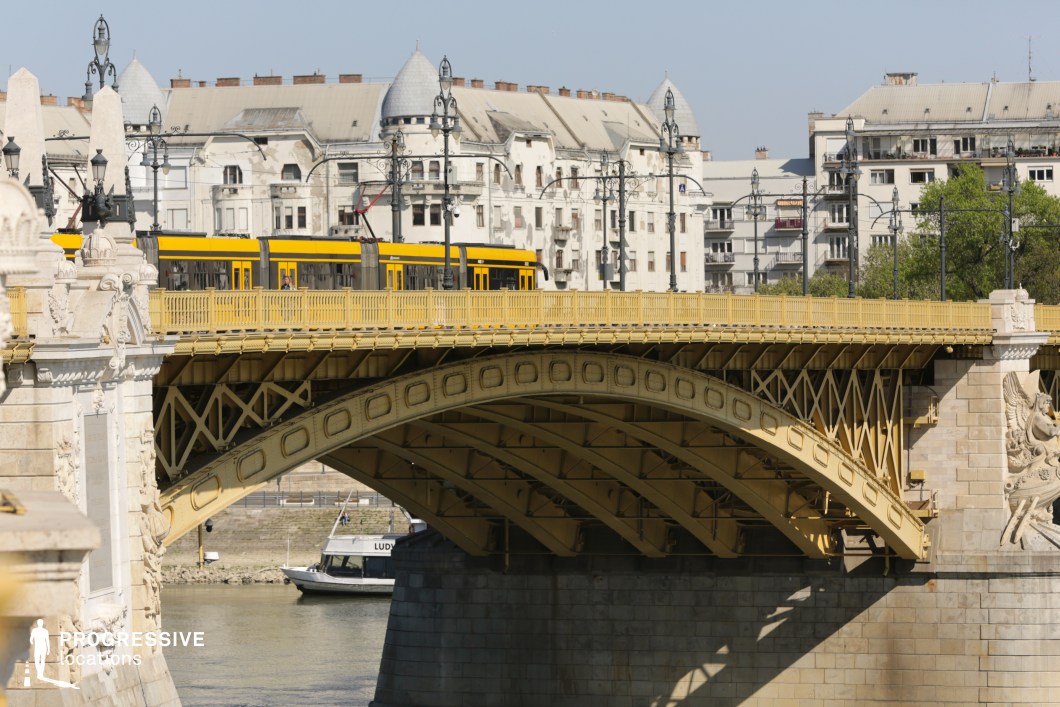 A striking view of an elegant metal bridge with intricate riveted arches, painted in a warm yellow hue, carrying a sleek modern tram across a river. The bridge is adorned with decorative lampposts and sculptural stone elements, blending classical and industrial design. In the background, historic residential buildings with domed rooftops and aged facades create a sense of timeless urban character. Below the bridge, a boat cruises along the river, adding to the dynamic cityscape. This visually rich setting is ideal for productions that seek a blend of historic charm and contemporary transportation infrastructure.