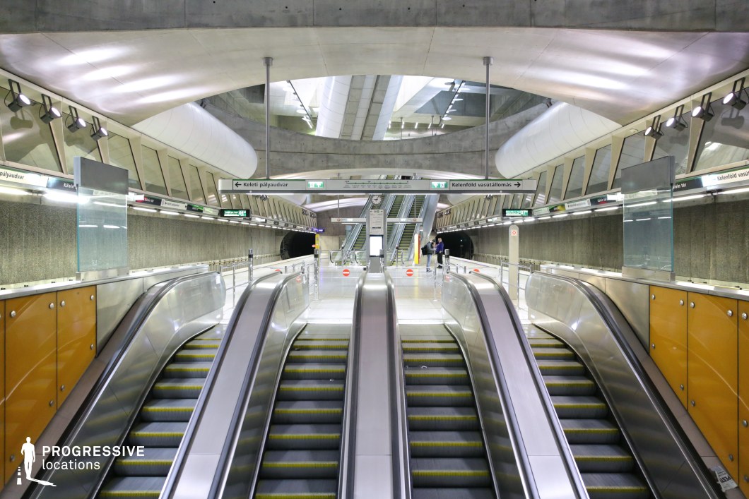 A sleek, ultra-modern metro station with an industrial aesthetic, featuring multiple escalators leading to an upper platform. The space is dominated by smooth concrete surfaces, reflective stainless steel, and geometric lighting elements. The curvilinear ceiling design adds depth and a futuristic feel, while signage provides directional guidance. The combination of polished floors, glass barriers, and structural beams creates a dynamic, high-tech atmosphere. This location is ideal for film productions seeking a contemporary urban transit setting with a clean, minimalist design.