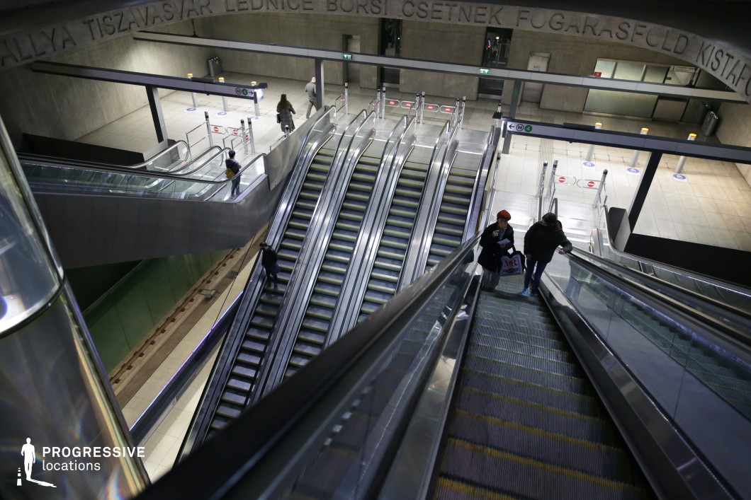 A vast and modern underground metro station featuring multiple escalators descending into a cavernous transit hub. The architecture is defined by raw concrete walls, stainless steel railings, and illuminated pathways, creating a sleek, industrial aesthetic. The large open space, combined with high ceilings and geometric design elements, conveys a sense of movement and scale. The station's signage and integrated lighting enhance its futuristic appeal, making it an ideal setting for scenes involving urban life, transportation sequences, or dystopian narratives.