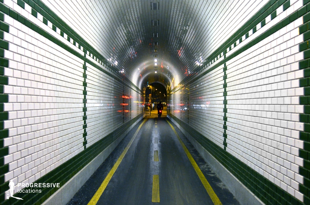 A narrow, arched tunnel lined with glossy white and green ceramic tiles, creating a striking geometric pattern. The tunnel is illuminated by overhead lights, which reflect off the tiles, enhancing the depth and symmetry of the space. The pathway features bold yellow lines, guiding movement towards the tunnel’s exit, where a cyclist is seen riding into the city night, framed by the tunnel’s curved opening. The tunnel’s reflective surfaces capture the ambient lights of the urban environment outside, adding a dynamic and almost futuristic quality to the scene. This setting is ideal for dramatic, cinematic sequences involving movement, transition, or urban exploration.
