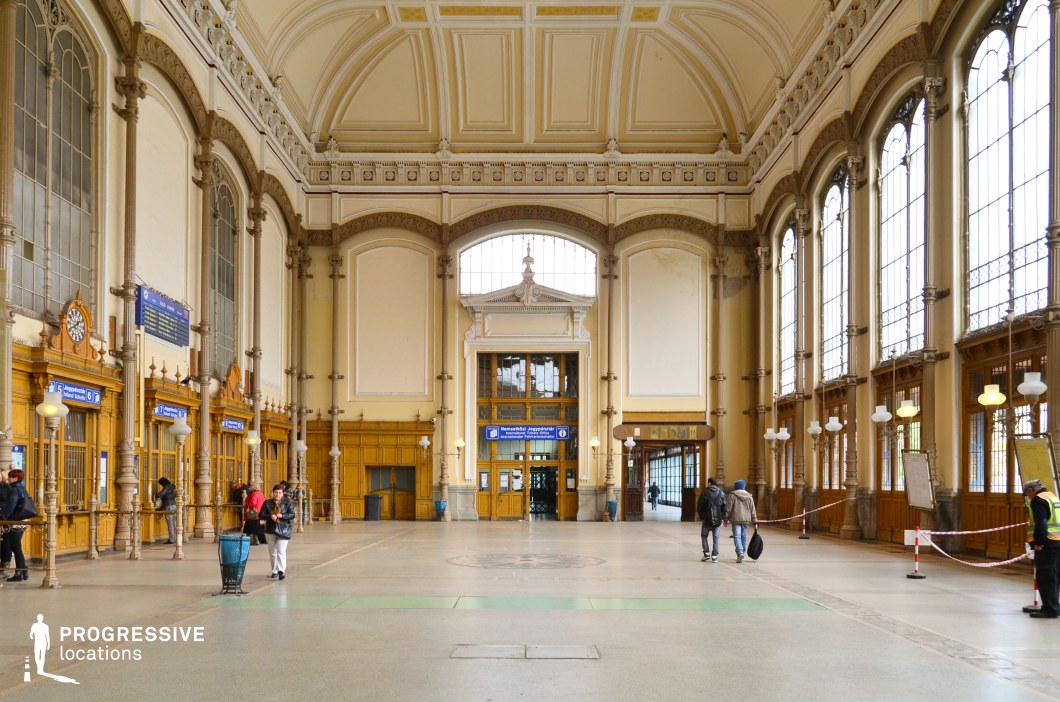 An opulent railway station ticket hall featuring grand neoclassical architecture, high vaulted ceilings, and intricate ornamental moldings. The symmetrical design is accentuated by towering arched windows that flood the space with natural light, casting a warm glow over the beige and cream-colored interior. Delicate decorative elements, including carved cornices and pilasters, add a sense of refinement and historic grandeur. The wooden ticket counters, framed by detailed wrought-iron railings and vintage-style signage, evoke a bygone era of railway travel. The polished stone floor, adorned with subtle geometric patterns, enhances the hall’s elegant and spacious atmosphere. Hanging globe lamps suspended from ornate brackets contribute to the classical aesthetic, complementing the rich wooden paneling along the walls. This grand yet functional space serves as a striking backdrop for period film productions, refined travel sequences, or architectural showcases, offering a timeless and visually compelling setting.