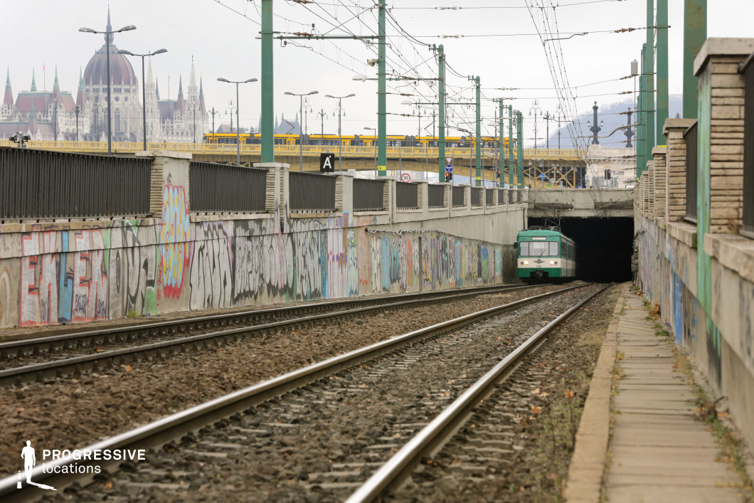 A gritty urban railway setting featuring a tunnel entrance framed by graffiti-covered concrete walls, contrasting with the ornate silhouette of a historic cityscape in the background. The converging train tracks create a strong sense of depth, leading the viewer’s eye toward the dark tunnel, where a green commuter train is emerging. Above the underpass, an elevated yellow tram crosses a bridge lined with decorative lampposts, adding a layer of vertical movement to the composition. The high walls flanking the tracks are adorned with colorful graffiti, lending the scene a raw, urban character. Overhead power lines crisscross the sky, reinforcing the industrial atmosphere, while the distant architectural landmarks provide an unexpected juxtaposition between modern transit infrastructure and classical European grandeur. This location presents a visually compelling mix of urban decay and historical elegance, making it ideal for cinematic cityscape shots, gritty crime narratives, or urban exploration photography.