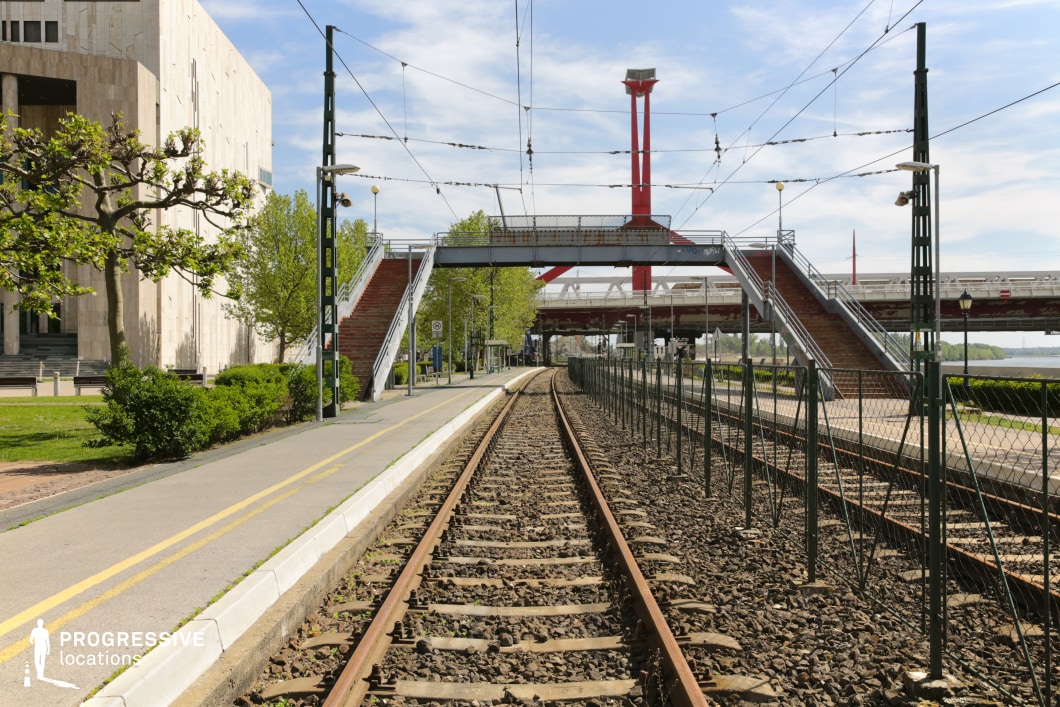 A contemporary railway setting featuring a long, straight tram track flanked by a pedestrian overpass and urban infrastructure. The tracks, embedded in gravel, extend into the distance, creating strong linear perspective lines that draw the eye towards the modern red bridge structure in the background. Overhead electrical wires crisscross the sky, supported by green metal poles, reinforcing the industrial transit aesthetic. The pedestrian bridge, with its symmetrical staircases and utilitarian design, provides safe access across the railway, adding depth and structural balance to the composition. To the left, a modern institutional building with a minimalist stone facade contrasts with the surrounding greenery, including neatly trimmed bushes and an elegantly sculpted tree. The right side is bordered by a metal fence, separating the tram line from a riverside area in the distance. This location blends urban transit elements with open space, making it an ideal backdrop for contemporary city scenes, transportation-focused storytelling, or architectural photography highlighting urban planning and infrastructure.